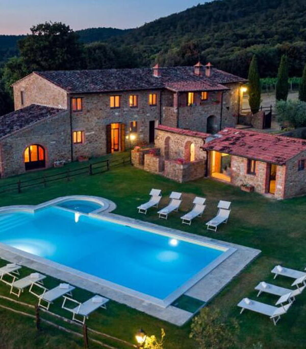Aerial evening view of an illuminated traditional stone farmhouse with an outdoor swimming pool, sun loungers, and lush green countryside at Castello di Meleto during a Baroque Bacchus tour