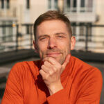 Smiling man with hand on chin wearing an orange sweater outdoors, sharing a positive review for a Baroque Bacchus tour in Tuscany