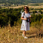 Young female traveler in a white dress, straw hat, and sunglasses holding a vintage camera on a hilltop overlooking Tuscan countryside during a Baroque Bacchus tour