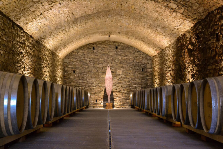 Symmetrical view of an ancient stone vaulted wine cellar lined with French oak barrels and featuring a modern central art sculpture at Castello di Meleto during a Baroque Bacchus tour