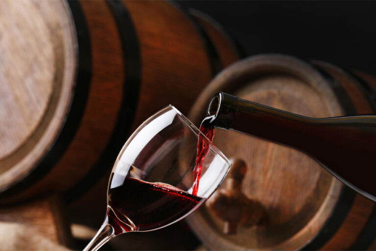 Red wine being poured from a bottle into a crystal glass with rustic wooden oak barrels in the background during a Baroque Bacchus wine tasting tour