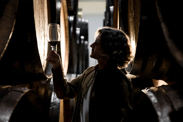 Sommelier or winemaker examining a glass of red wine against the light in a dark cellar surrounded by large oak aging barrels during a Baroque Bacchus tour