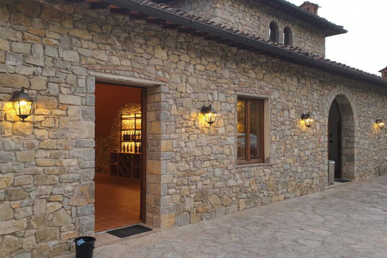 Rustic stone building of an authentic Tuscan winery estate with warm lanterns lit at dusk, featuring an open doorway showing a wine display inside