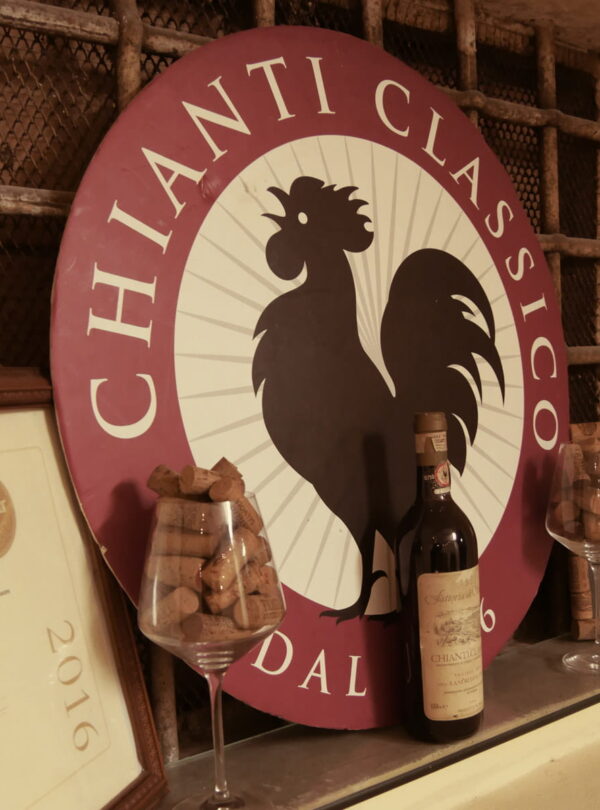 Iconic circular Chianti Classico sign featuring the Black Rooster logo alongside a bottle of Chianti Classico wine, wine corks, and award certificates during a Baroque Bacchus tour